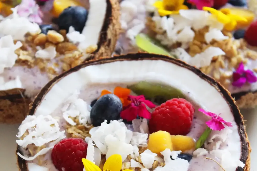 Bowl aus einer halben Kokosnuss mit Beeren, Mango, Kokosraspeln und essbaren Blüten