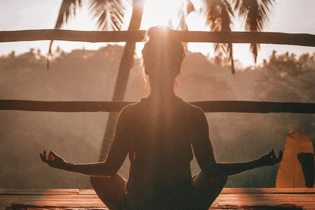 Person in Meditationshaltung auf einer Holzterrasse, im Gegenlicht der untergehenden Sonne zwischen Palmen
