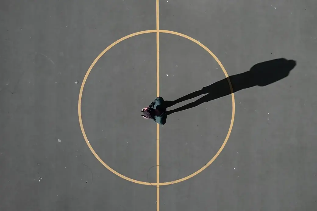 A person in the centre circle of a sports court, seen from above, casting a long shadow
